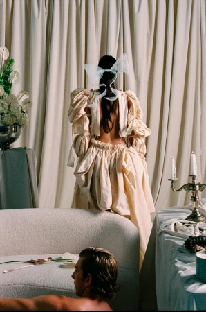 Woman in a cream dress with puffy sleeves stands in front of draped curtains; a man lies on a white couch in the foreground.