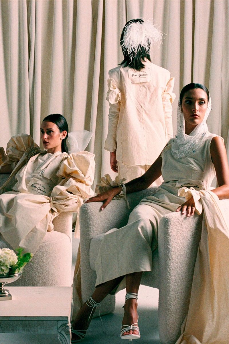 Three elegant women in stylish, cream-colored dresses pose in a bright indoor studio setting with soft lighting and elegant furniture.