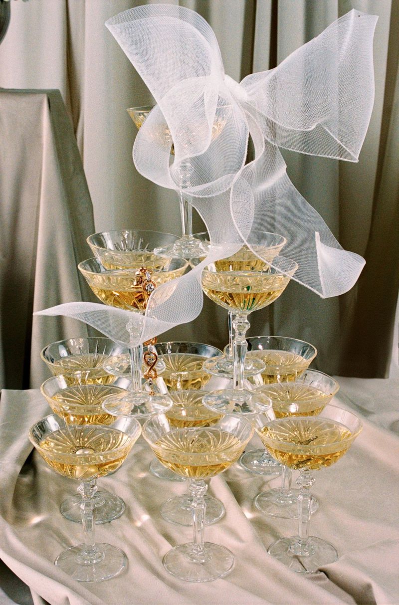 Champagne tower with stacked crystal coupe glasses filled with golden liquid, decorated with a white mesh bow in an indoor setting.