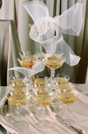 Champagne tower with stacked crystal coupe glasses filled with golden liquid, decorated with a white mesh bow in an indoor setting.