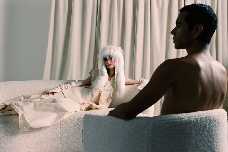 Fashion photo of a woman with a shaggy white wig lying on a sofa as a man with a bare chest sits in foreground chair.