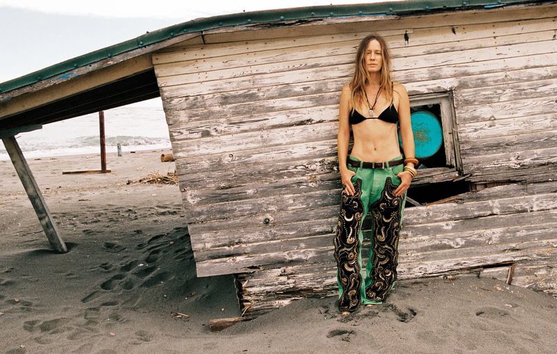 Model Nataša Vojnović wearing a black bikini top and green embroidered trousers stands beside a weathered wooden beach shack.