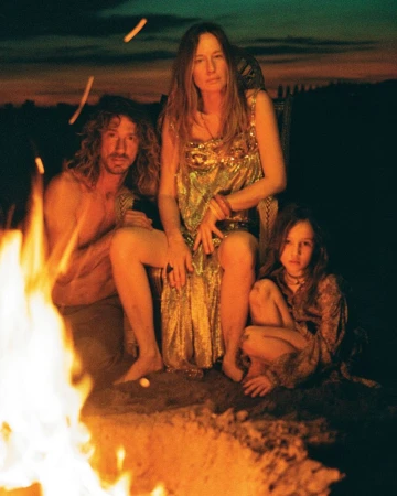 Model Nataša Vojnović in a gold dress sitting beside a man and a child near a glowing campfire at dusk.
