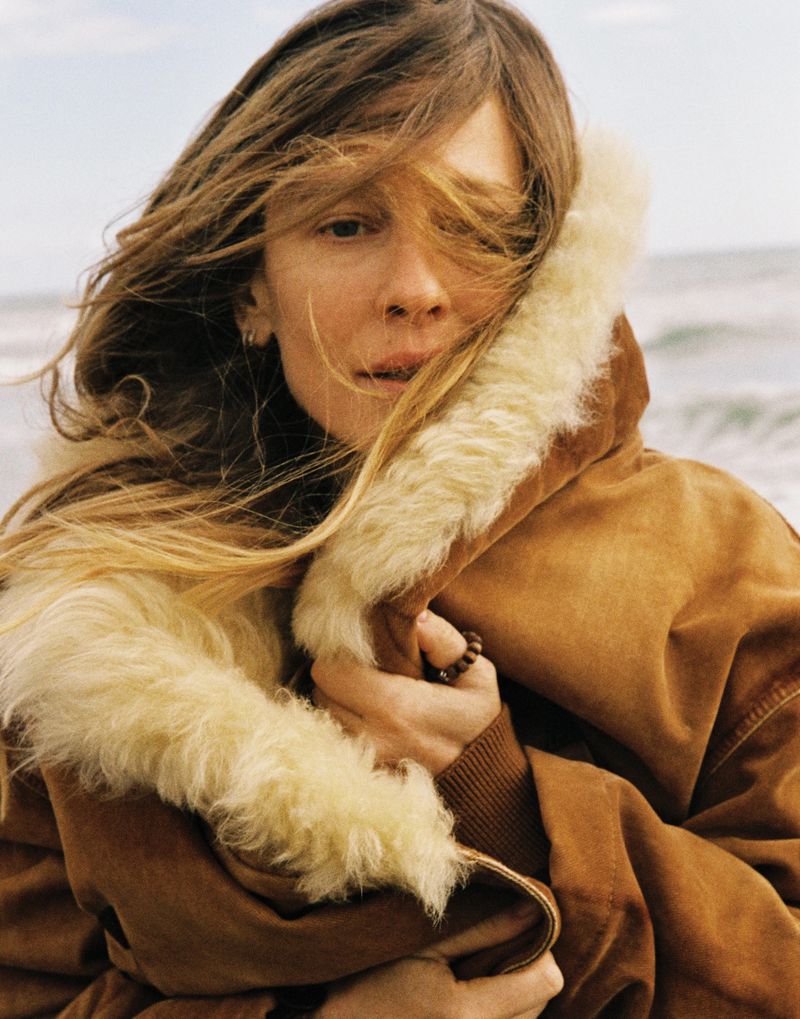 Model Nataša Vojnović wrapped in a brown shearling coat with windblown hair against a coastal ocean backdrop.