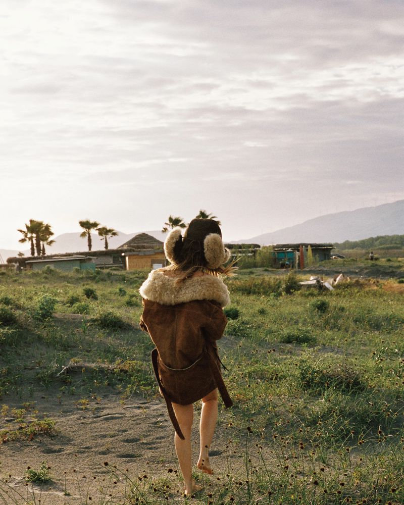 Model Nataša Vojnović walks away barefoot across a grassy sand landscape wearing a belted brown shearling coat and plush earmuffs.