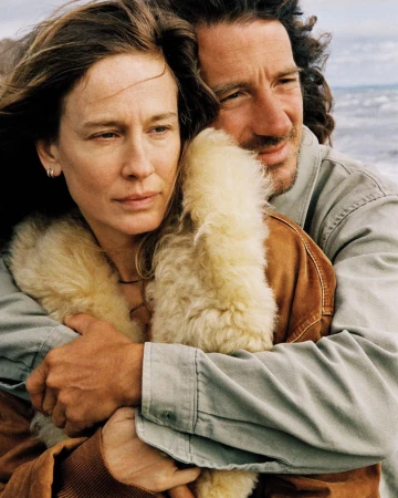 Close-up of Nataša Vojnović wearing a brown shearling coat embraced from behind by a man outdoors by the coast.