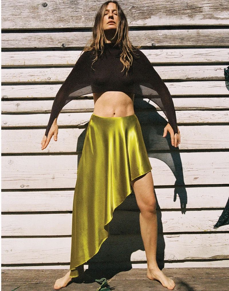 Nataša Vojnović poses barefoot in a black batwing crop top and chartreuse asymmetric satin skirt against a wooden wall.