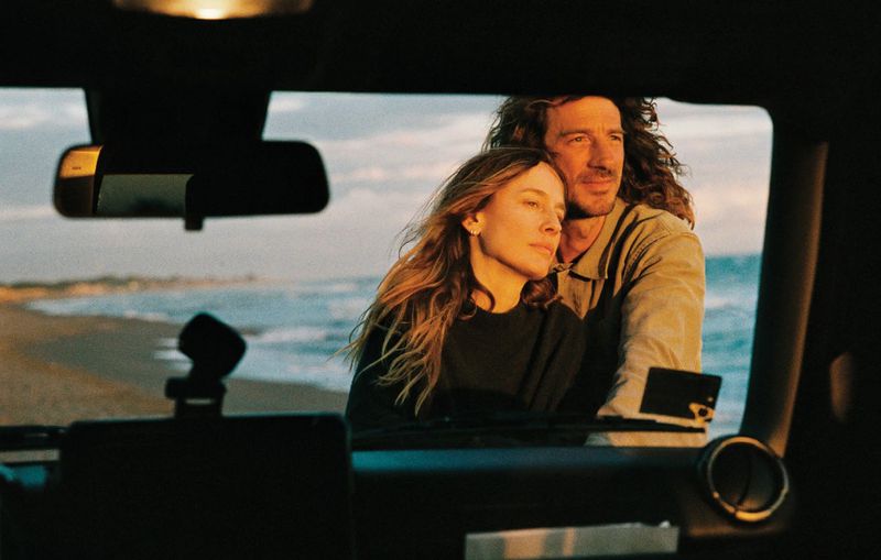 Natasa Vojnovic and a man seen through a car windshield standing on a sandy beach at sunset.