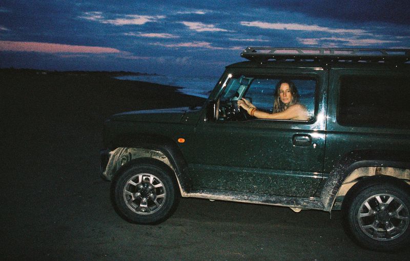 Model Nataša Vojnović seated inside a dark green off-road vehicle parked on a beach at dusk.