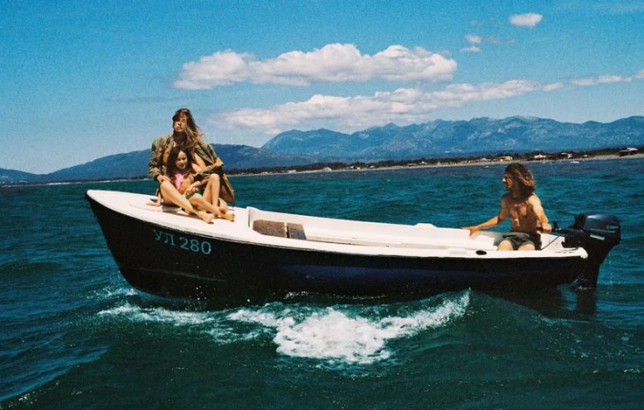 Nataša Vojnović and a child sit on the bow of a small motorboat navigated by a man on open blue water against distant mountains.
