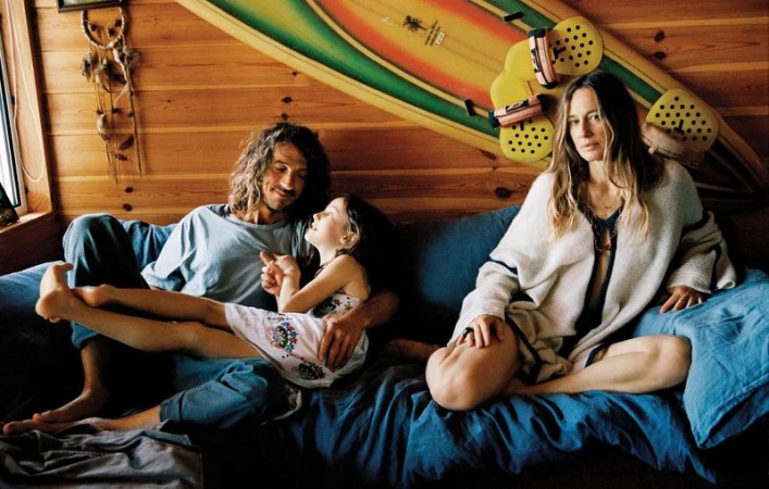 Natasa Vojnovic sitting on a blue sofa beside a man and young girl against a wood-paneled wall with a surfboard.