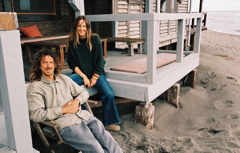 Nataša Vojnović in a green sweater and blue jeans sitting on a wooden beach shack porch alongside a man in a folding chair.