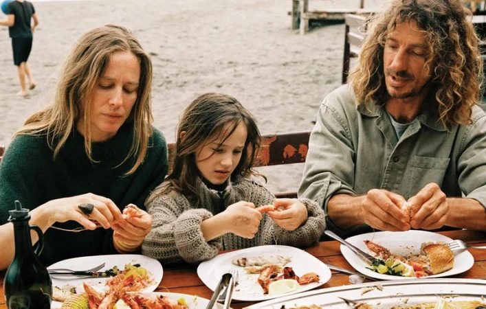 Nataša Vojnović, a young girl, and a man seated at a wooden outdoor table peeling seafood on a sandy beach.