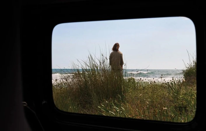 A view through a dark vehicle window of model Nataša Vojnović standing among tall coastal dune grass facing the sea.