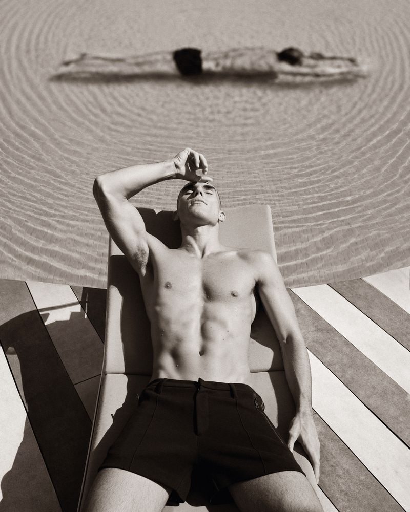 A shirtless man in black shorts relaxes on a sun lounger beside a swimming pool with another swimmer underwater in the background.