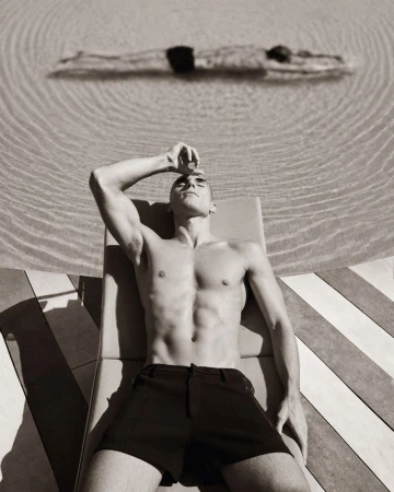 A shirtless man in black shorts relaxes on a sun lounger beside a swimming pool with another swimmer underwater in the background.
