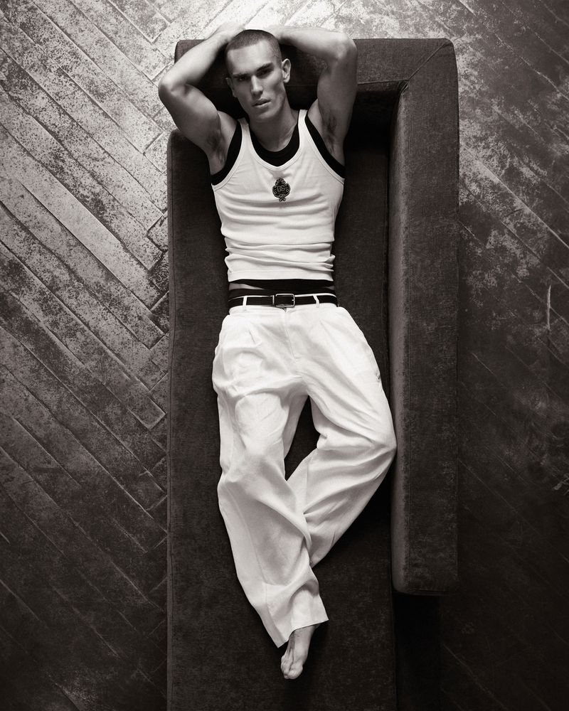 Model Josh McGregor lying barefoot on a couch wearing a white ribbed tank top and white trousers on parquet flooring.