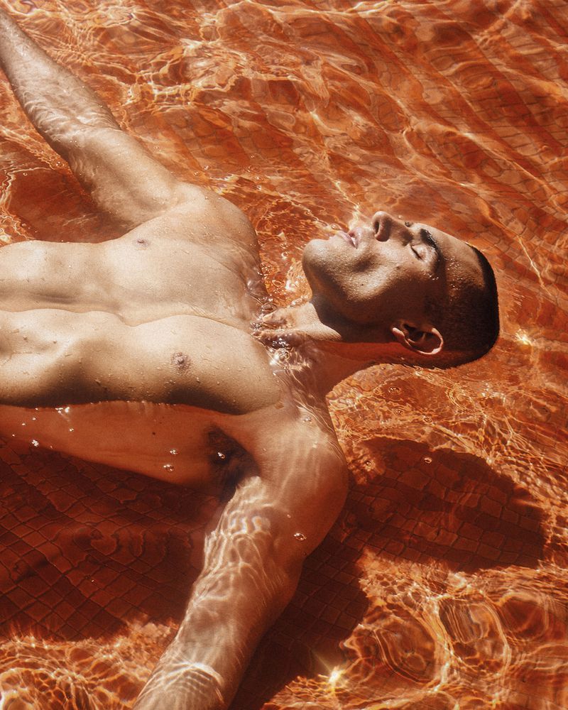 Model Josh McGregor floating on his back in shallow pool water with rippling sunlight caustics over tiled pool floor.