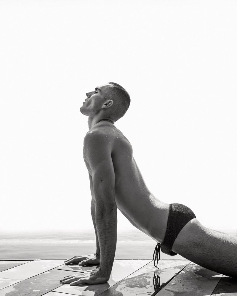 Model Josh McGregor performing an upward stretch in dark swim briefs on a wet poolside deck in black and white.