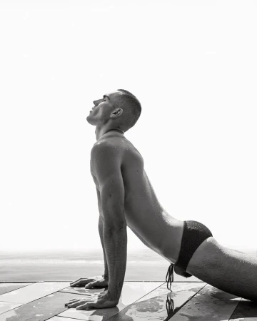 Model Josh McGregor performing an upward stretch in dark swim briefs on a wet poolside deck in black and white.
