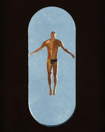 Model Josh McGregor in swim trunks floating over clear blue water viewed through an elongated oval aperture on a black ground.