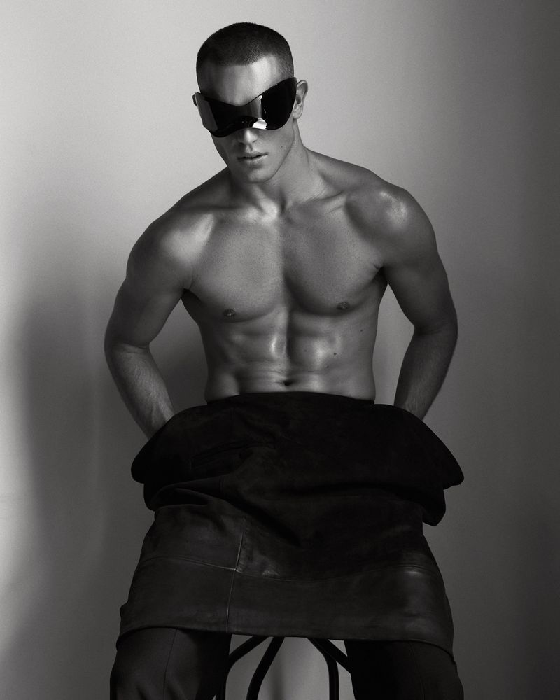 Shirtless male model wearing futuristic black shield sunglasses seated against a neutral studio backdrop.