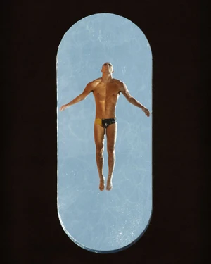Model Josh McGregor in swim trunks floating over clear blue water viewed through an elongated oval aperture on a black ground.