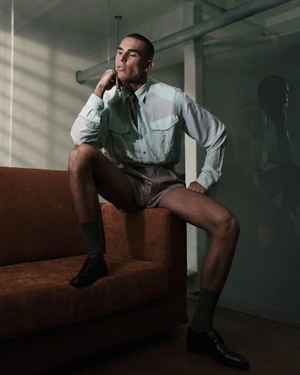 Model Josh McGregor seated on the arm of a sofa wearing a pale mint shirt, tailored shorts, and black shoes in a shadowed room.