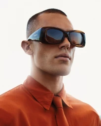 Josh McGregor wearing oversized brown sunglasses and a rust-orange collared shirt with matching tie against a bright sky.