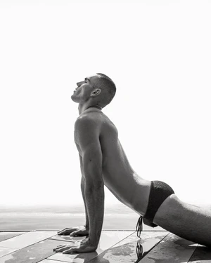Model Josh McGregor performing an upward stretch in dark swim briefs on a wet poolside deck in black and white.