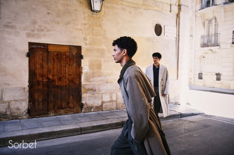A man in profile wearing an oversized taupe trench coat walks along a city street with another model in a beige coat behind him.