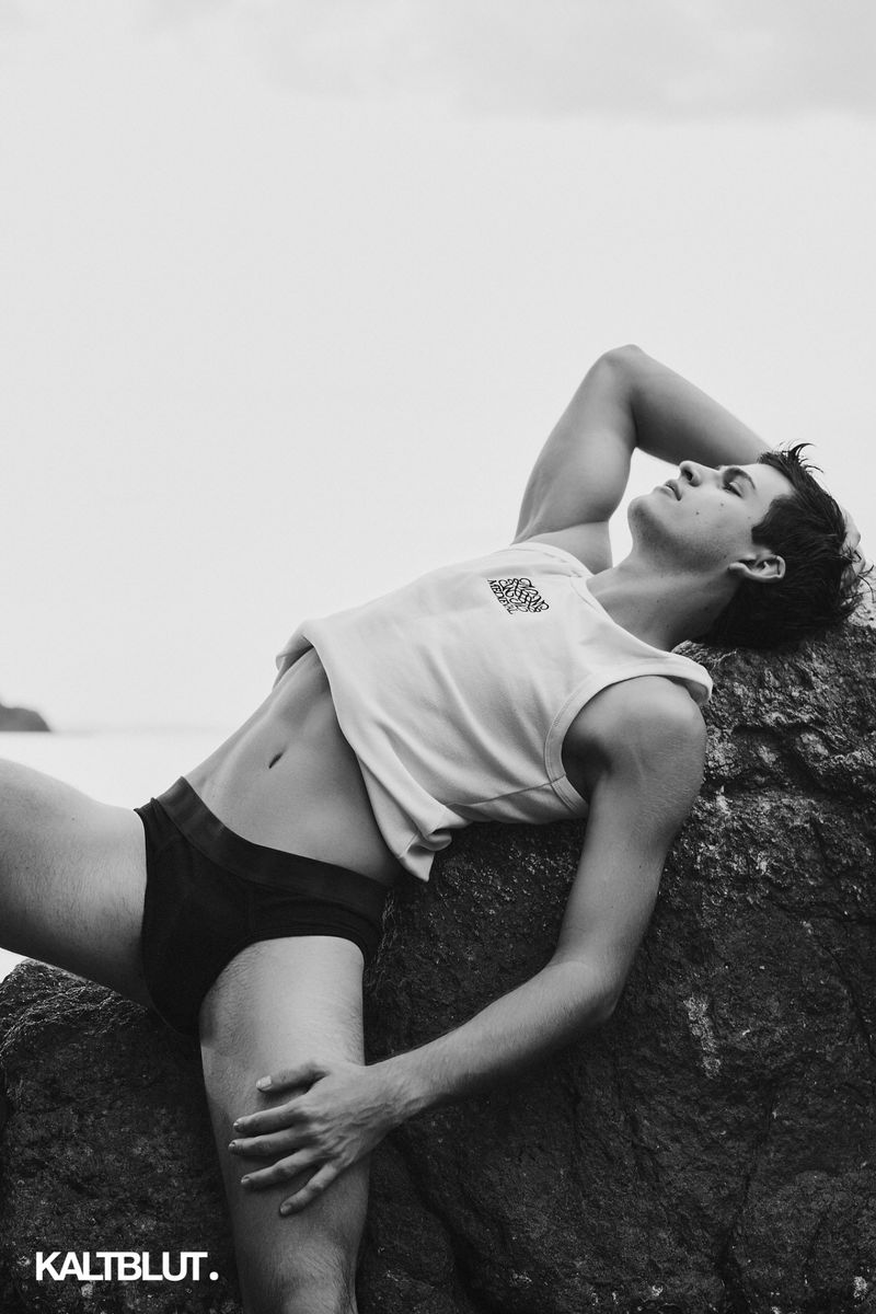 Man in a white sleeveless crop top and black briefs reclining on rocky coastal boulders in black and white.