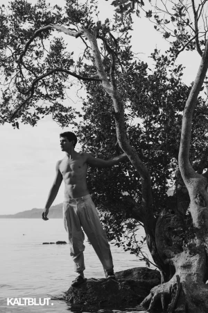 Shirtless man standing on rocks by the water beneath a twisted tree branch for editorial shoot