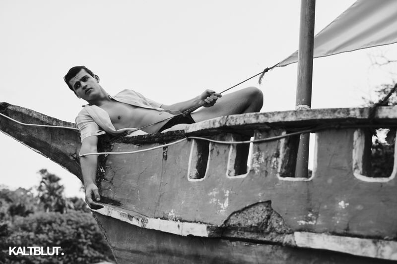 Young man reclining on the bow of a wooden boat, holding a rope against a pale sky and foliage background.