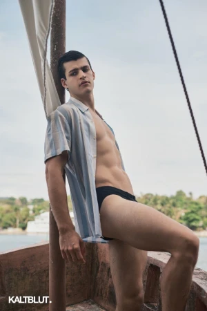 A man posing on a wooden boat wearing an unbuttoned blue striped shirt and black swim briefs for Kaltblut Magazine.