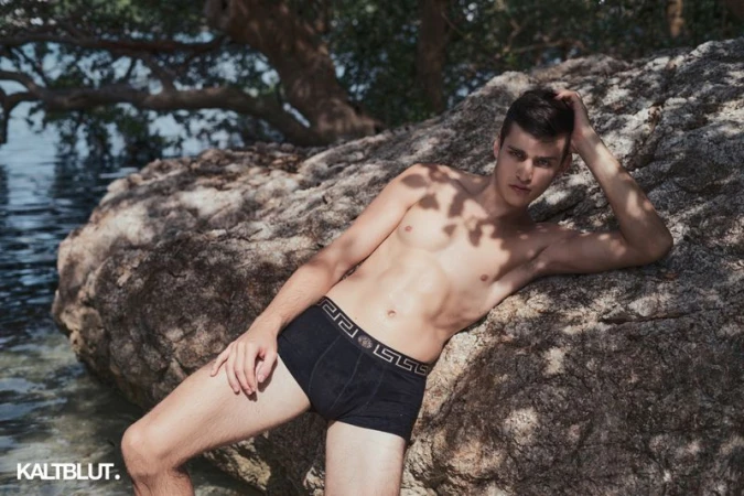 A male model wearing black patterned boxer briefs reclines against a large rocky boulder by coastal water in dappled sunlight.