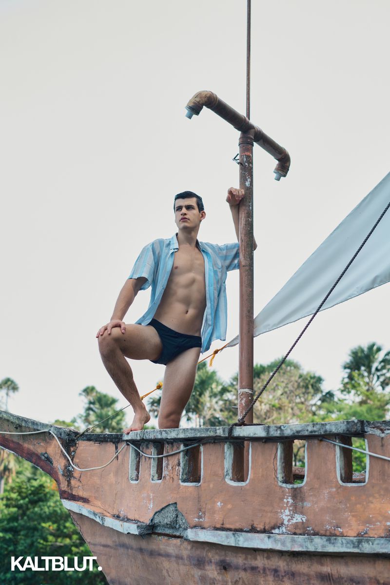 A man in an open light blue shirt and dark swim briefs poses on the bow of a weathered boat under an overcast sky.