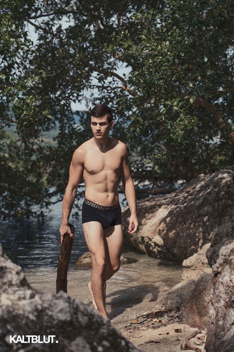 Shirtless man in black swim trunks carrying driftwood along a rocky coastline under tree branches.