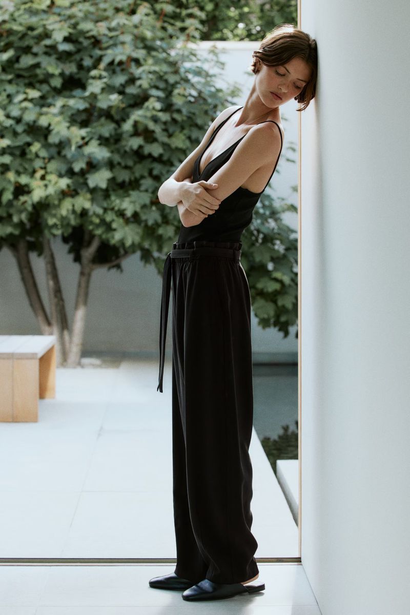 Model Valerie Scherzinger leaning against an interior wall wearing a black sleeveless top and loose black trousers.