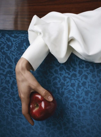 Close-up of a hand in a voluminous white sleeve holding a glossy red apple against patterned blue fabric and polished wood.