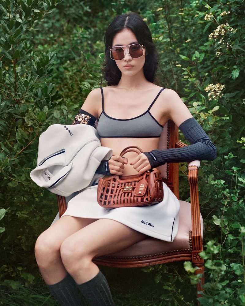 Young woman in outdoor fashion shot wearing gray bra top, white skirt, knee socks, carrying brown purse and white jacket by Miu Miu.