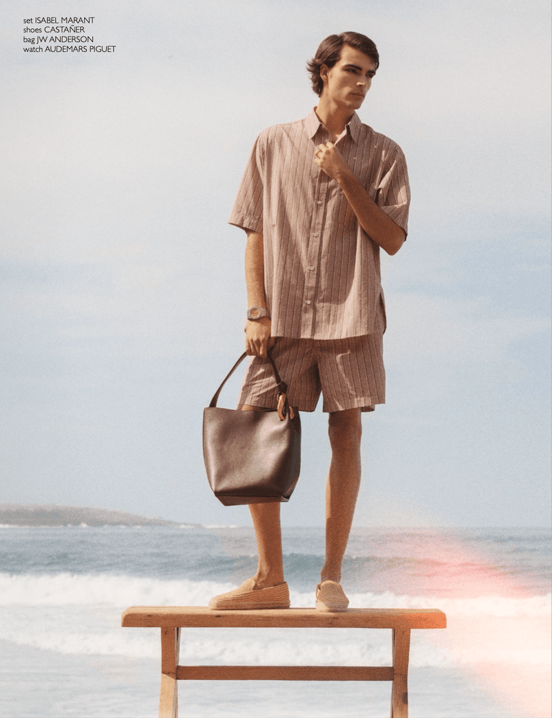 A young man standing on a wooden bench by the sea, wearing a striped shirt and shorts, holding a brown leather shoulder bag.