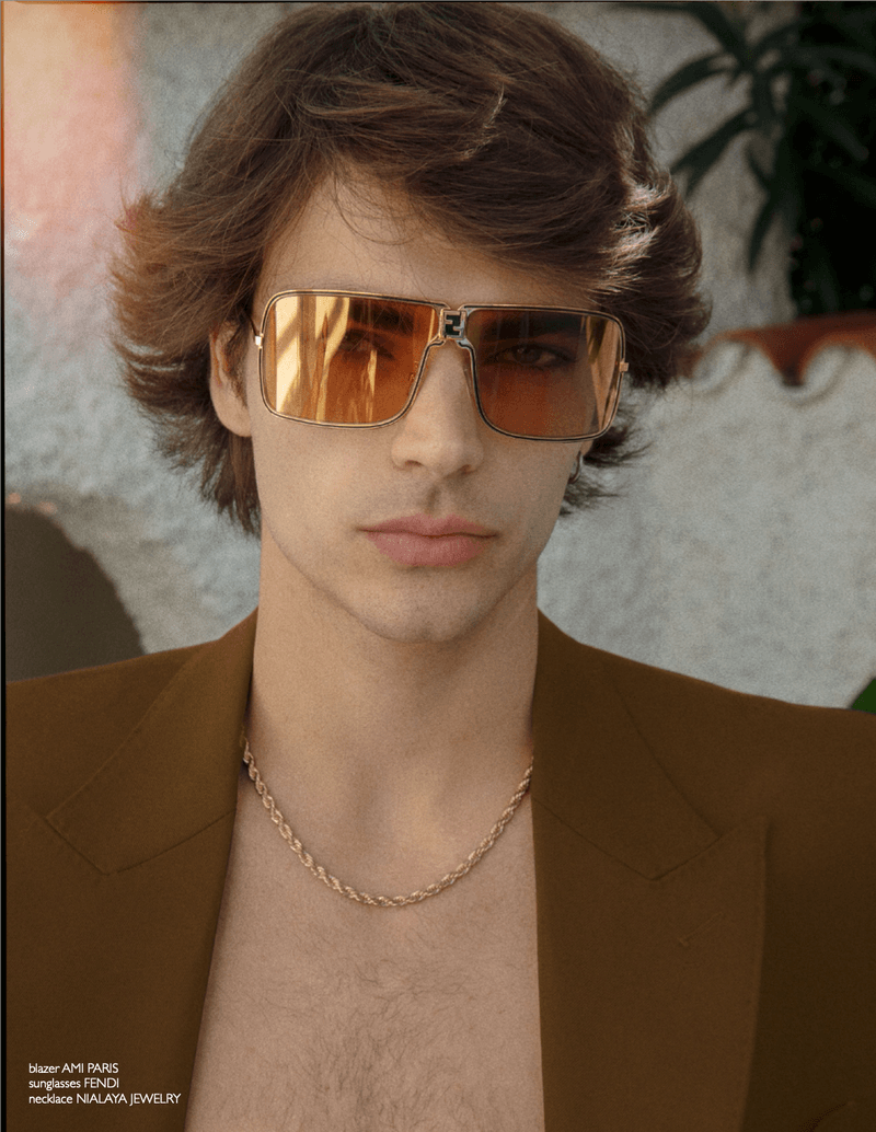 Model Rafa Noah wearing tinted square sunglasses, a gold rope chain, and an open brown blazer in a close-up portrait.