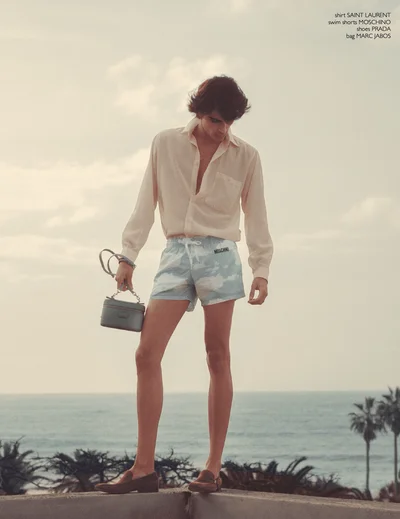 Model standing on a ledge overlooking the ocean in a cream button-up shirt, printed swim shorts, loafers, and a handbag.