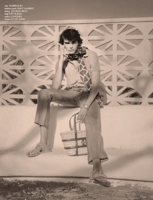 Model Rafa Noah seated on a ledge wearing a polo shirt, patterned neck scarf, and sandals with a basket tote bag beside him.