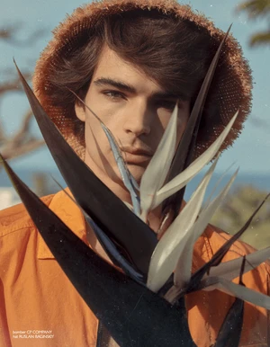 Model Rafa Noah in a textured straw hat and orange jacket holds a bird of paradise flower in front of his face.