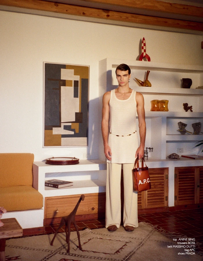 Model Rafa Noah wearing a long cream tank tunic and wide-leg trousers holding a brown leather bag in a styled living room.
