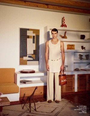 Model Rafa Noah wearing a long cream tank tunic and wide-leg trousers holding a brown leather bag in a styled living room.