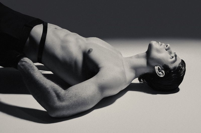 Black and white portrait of shirtless model Niccolo Fortini reclining backward on the floor with strong directional shadows.