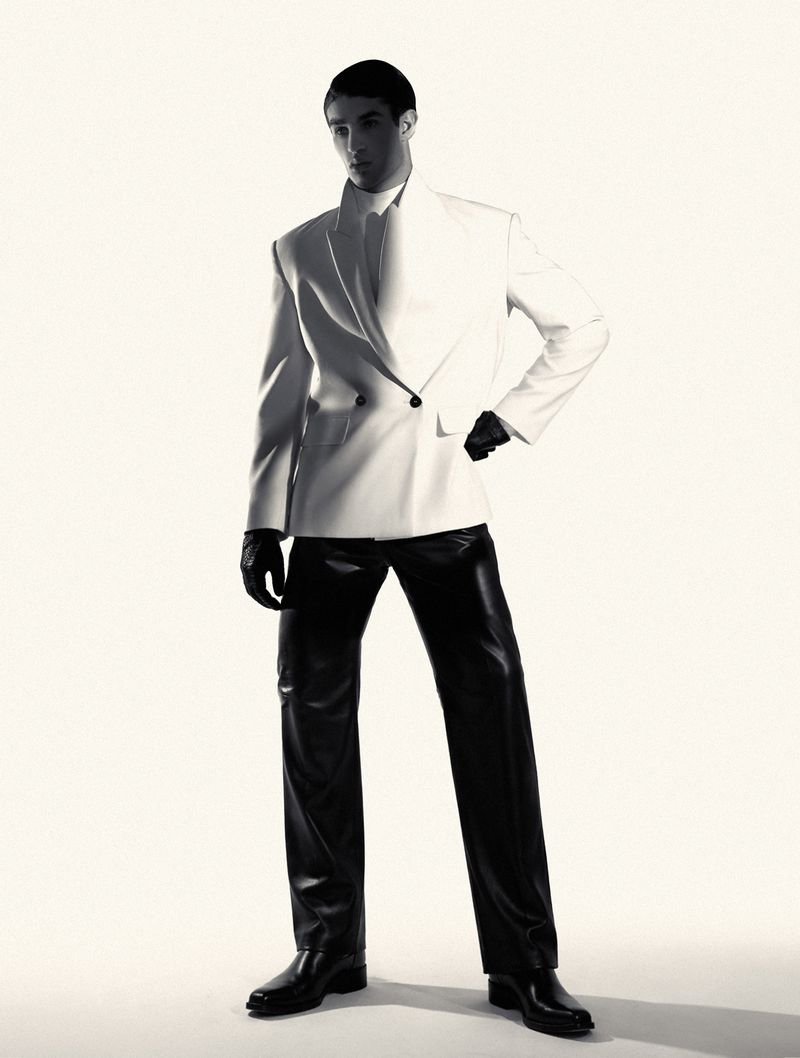Model in a structured white double-breasted blazer, black leather pants, and gloves standing against a white studio background.
